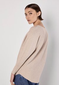 Beige pletený svetr s oversized střihem, kulatým výstřihem a bočními rozparky, v kombinaci s modrými džíny. Hladká textura a uvolněný tvar.