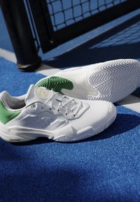 Scarpe da ginnastica bianche e verdi posizionate su un campo sportivo blu vicino a un palo della rete nero e una linea di confine bianca.