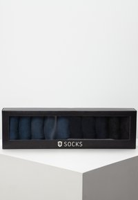 Ensemble de huit chaussettes dans des nuances variées de bleu marine et de noir, présentées dans une boîte rectangulaire noire avec un panneau avant en verre transparent.