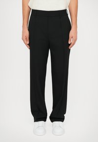 Black trousers with a relaxed fit, smooth fabric, and pleats. Paired with white sneakers, featuring a classic design and straight leg.