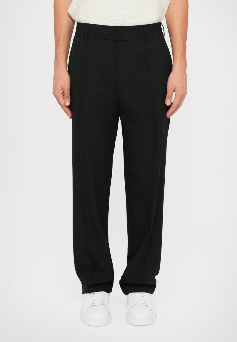 Black trousers with a relaxed fit, smooth fabric, and pleats. Paired with white sneakers, featuring a classic design and straight leg.