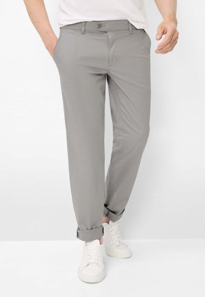 Uomo che indossa pantaloni grigi chiari risvoltati, sneakers bianche e una camicia bianca con le mani nelle tasche, in piedi davanti a uno sfondo bianco.