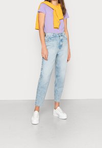 Ljusblå högmidjade jeans med rak benpassform, kombinerat med en kort lila t-shirt och en gul sweatshirt hängande över axlarna.