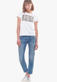 Balta kokvilnas T-kreklu ar rakstainu melnbaltu grafik un nolietiem zili džinsiem, kā arī baltiem sporta apaviem.