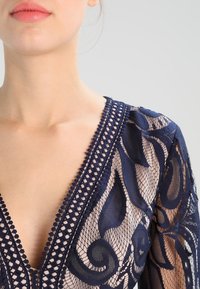 Marineblaues Spitzenoberteil mit filigranen Blumenmustern, tiefem V-Ausschnitt und strukturierter Stickerei entlang des Ausschnitts und der Ärmel.