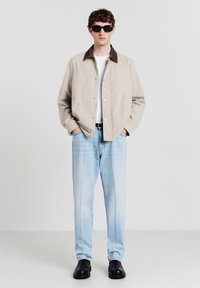 Veste beige avec col marron, chemise blanche, jean bleu clair et chaussures noires. Coupe slim avec fermeture à boutons et design minimal.