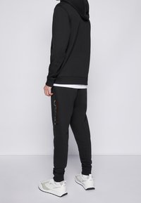 Felpa nera con cappuccio regolabile, abbinata a pantaloni grigio scuro con stampa logo lucida e sneaker bianche con dettagli neri.