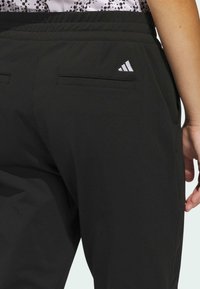 Svarta sportbyxor med elastisk midjeresår, två bakfickor och en liten vit Adidas-logotyp på övre högra sidan. Slätt, lättviktigt tyg.