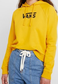 Felpa gialla con un logo nero "VANS", dotata di cappuccio con lacci, polsini a coste e un fit morbido, abbinata a jeans in denim a vita alta.