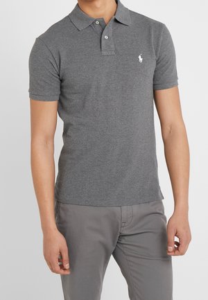 Homme portant un polo gris ajusté à manches courtes avec deux boutons et un pantalon gris, debout devant un fond uni.