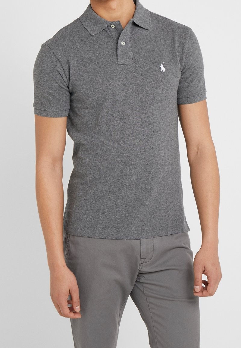 Homme portant un polo gris ajusté à manches courtes avec deux boutons et un pantalon gris, debout devant un fond uni.