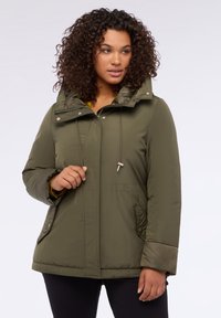 Oliva vihreä pehmustettu takki hupulla, jossa on nyörityksellä varustettu yksityiskohta ja sivutaskut. Suunnittelussa on vetoketjukiinnitys ja sileä, teksturoitu kangas.