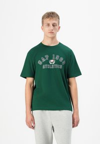Grønn bomull T-skjorte med "GAP 1969 ATHLETICS" i hvite og røde bokstaver. Designet med rund hals og korte ermer.