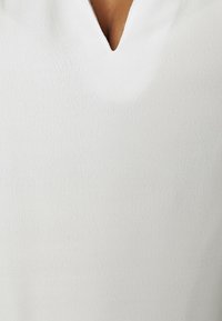 Vit blus med V-ringning, gjord av ett mjukt tyg med en subtil textur. Designen är enkel utan ytterligare dekorationer.