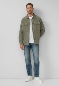 Olive green corduroy jacket z zapięciem na guziki, z dwiema kieszeniami na piersi, noszona na białym t-shircie, zestawiona z jasnoniebieskimi dżinsami i białymi trampkami.