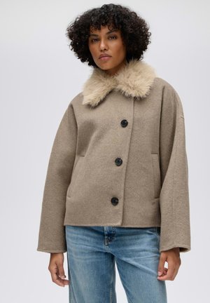 Donna con capelli ricci che indossa un cappotto di lana beige con colletto soffice e bottoni neri, abbinato a jeans blu, in piedi davanti a uno sfondo semplice.