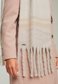 Beige uldsjael med frynsede kanter, prydet med subtile lyse orange striber. Sjaelen hænger over en pink frakkeslange, hvor et metallisk logo er synligt.