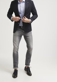 Blazer blu navy sopra una camicia azzurro chiaro, abbinato a jeans grigi attillati e scarpe eleganti nere. Caratteristiche includono una tasca sul petto e maniche arrotolate.