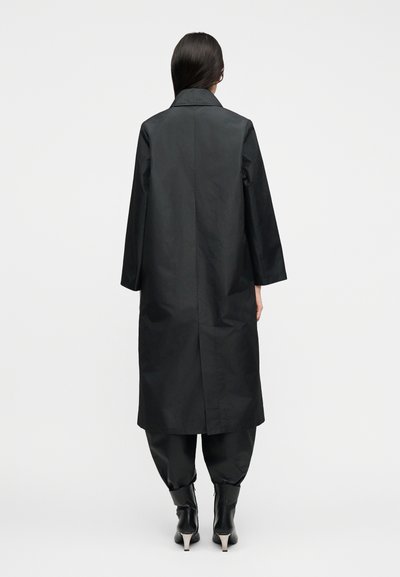 Γυναίκα με μακριά σκούρα μαλλιά, ντυμένη με μαύρο μακρύ παλτό, μαύρο στενό παντελόνι και μαύρα ψηλοτάκουνα μποτάκια, στέκεται μπροστά σε απλό φόντο.