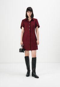 Donna in un vestito bordeaux bottonato fino al ginocchio, con una piccola borsa nera in mano e stivali neri fino al ginocchio, in piedi davanti a uno sfondo bianco.