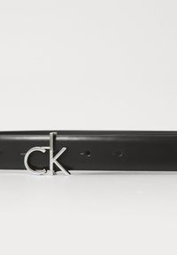 Svart lærbelte med en slank sølv "CK" logo spenne, med glatt tekstur og flere hull for størrelsesjustering.