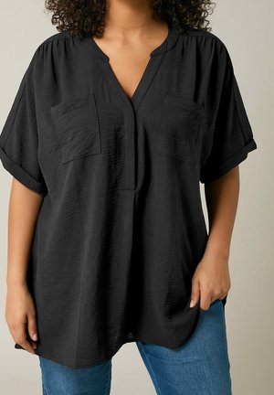 Kvinde iført løs sort tekstureret kortærmet bluse med to brystlommer, kombineret med blå jeans mod en ensfarvet baggrund.