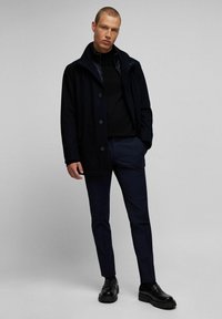 Marineblauwe jas met een hoge kraag, vijf knopen, gelaagd over een zwarte coltrui en gecombineerd met donkere broek en stevige zwarte schoenen.