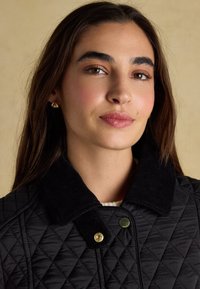 Femme aux longs cheveux foncés porte une veste matelassée noire avec un col en velours côtelé et des boutons-pression dorés, fond neutre.