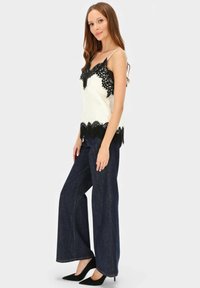 Biała koszulka na ramiączkach ozdobiona czarną koronką, zestawiona z szerokimi spodniami z ciemnego denimu. Modelka nosi czarne szpilki w szpic, włosy opadają jej na ramiona.