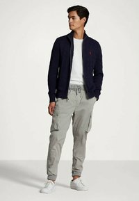 Ung man som står, iklädd en marinblå zip-up-tröja, vit t-shirt, ljusgrå cargobyxor och vita sneakers, som tittar åt vänster.