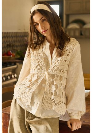 Mladića žena u krem čipkastoj bluzi i prsluku, s odgovarajućom trakom za glavu, naslonjena na drveni stol u ugodnoj kuhinji.