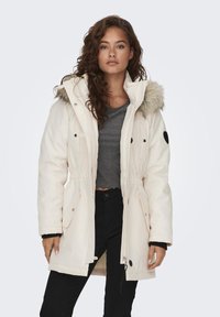 Witte parka met een met bont gevoerde capuchon, een getailleerde taille, zwarte accenten en meerdere zakken. Draag het over een grijs topje en zwarte broek.