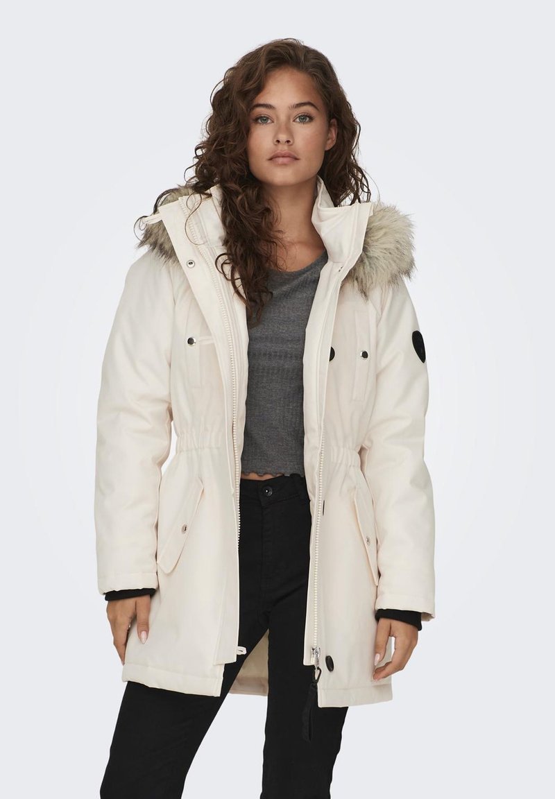 Witte parka met een met bont gevoerde capuchon, een getailleerde taille, zwarte accenten en meerdere zakken. Draag het over een grijs topje en zwarte broek.