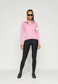 Maillot de cyclisme rose à manches longues avec fermeture éclair, bande blanche sur la manche, associé à un collant de cyclisme noir. Le porteur se tient avec confiance dans des chaussures de cyclisme.