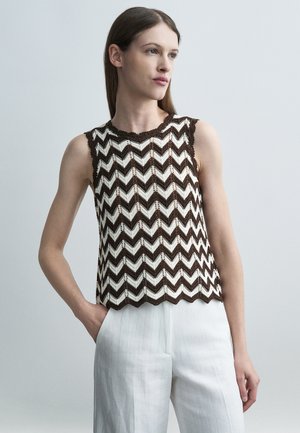 Γυναίκα που φορά αμάνικο καφέ και λευκό τοπ με σχέδιο ζιγκ-ζαγκ και λευκό παντελόνι, στέκεται με το ένα χέρι στην τσέπη.