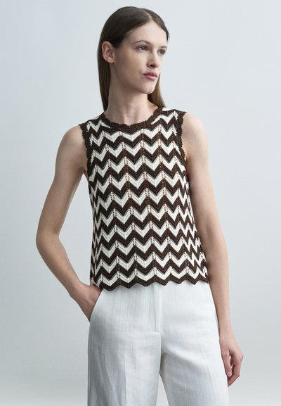 Femme portant un haut sans manches à motif chevron marron et blanc et un pantalon blanc, debout avec une main dans la poche.