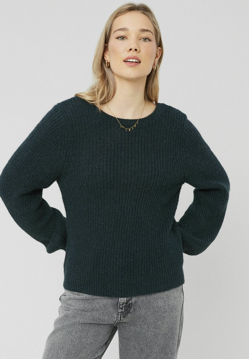 Pull en tricot vert foncé avec un col rond, des manches longues et un design à côtes texturé, associé à un jean gris clair.