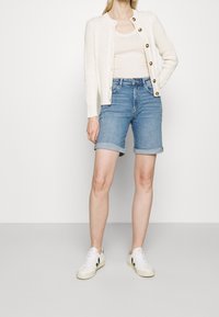 Cremefarbener gestrickter Cardigan mit goldenen Knöpfen, beigem Tanktop und hochgekrempelten blauen Jeansshorts. Weiße Sneakers mit schwarzen Akzenten.