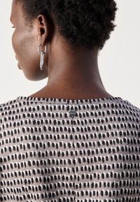 Blouse à motifs avec une base grise et des points noirs et blancs, un col rond et une fermeture à boutons au dos.