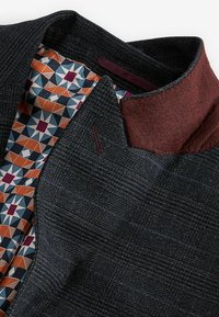 Jachetă cu model închis, având un exterior texturat și o căptușeală cu model geometric în nuanțe de portocaliu, albastru și crem; detalii în nuanță de vișiniu la guler.