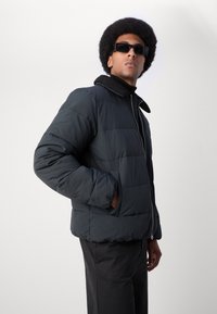 Mørk polstret jakke med lynlås foran, lammeskindsfarvet krave og sidelommer, lavet af glat stof. Modellen har mørke solbriller og bukser på.