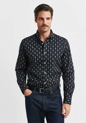 Mann mit Schnurrbart, der ein marineblaues, paisleygemustertes Hemd trägt, das in eine dunkelblaue Jeans mit schwarzem Gürtel gesteckt ist, und vor einem einfarbigen Hintergrund steht.