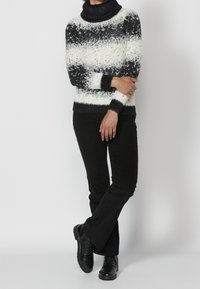 Person, der einen schwarz-weißen gestreiften Rollkragenpullover, schwarze Hose und schwarze Schnürstiefel trägt und mit verschränkten Armen dasteht.