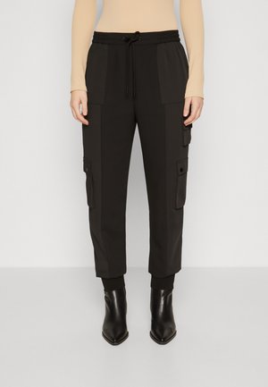 Musta vetoketjullinen vyötärönyörilliset cargo-housut, sivutaskuilla ja käännetyillä lahkeilla, yhdistetty beigeen pitkähihainen puseroon ja mustiin teräväkärkisiin nilkkureihin.