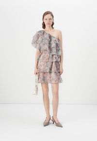 Kvinna står klädd i en blommig enaxlad klänning med lager av volanger, hållande en vit handväska, beige slingback-klackar, mot en enkel vit bakgrund.