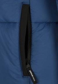 Blau quadratisch gepolsterte Jacke mit sichtbar gezipptem Reißverschluss, schwarzem Innenfutter und Etikett mit Markenname. Die Textur wirkt glatt und strapazierfähig.
