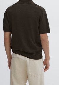 Personne portant un pull à manches courtes marron foncé et un pantalon beige clair, vue de dos sur un fond uni.