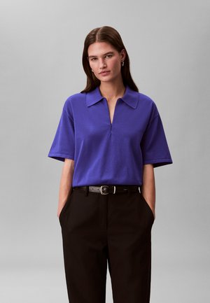 Jeune femme aux cheveux bruns et raides portant un polo à manches courtes violet rentré dans un pantalon noir avec une ceinture noire, les mains dans les poches.