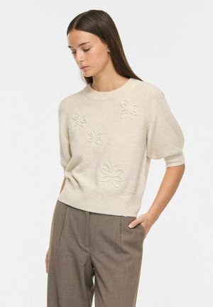 Giovane donna che indossa un maglione crema a maniche corte con fiori ricamati e pantaloni marroni a vita alta, guarda verso il basso con una mano in tasca.