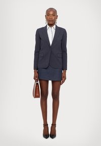 Blazer blu scuro con rever a punta, abbinato a una gonna di jeans. Il modello tiene una borsa marrone. Scarpe nere con tacco alto completano l'outfit.